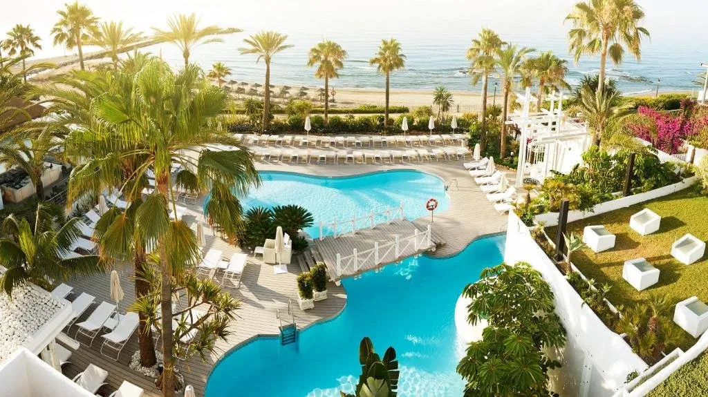 Pools among tall palms at Puente Romano, crossed by a timber footbridge, with white loungers on the decking and the beach and the sea beyond