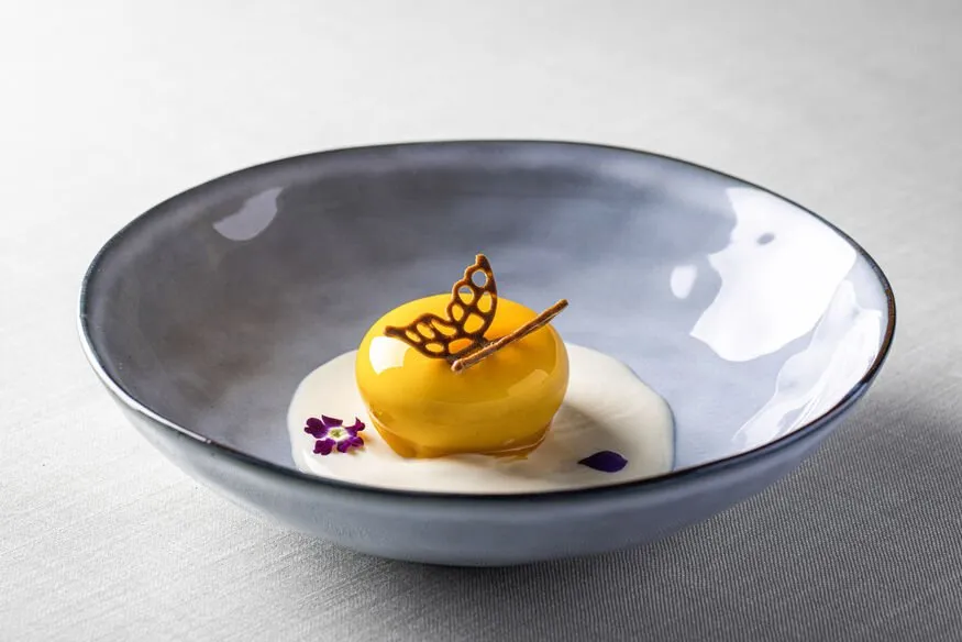 A glossy yellow dome crowned with a filigree butterfly, on a pool of white cream in a grey bowl, with a small violet flower