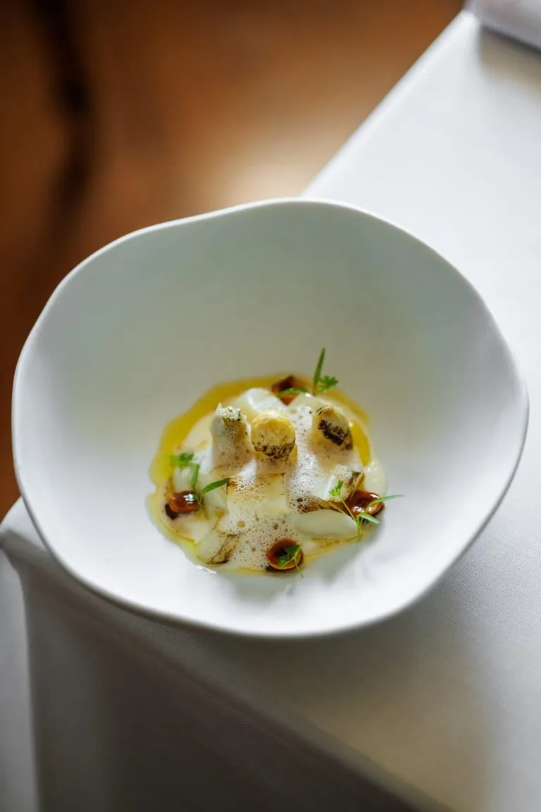 A white bowl with a wavy rim on a white tablecloth, holding pale grilled vegetables under a light foam, with a drizzle of oil, small toasted pieces and sprigs of herbs