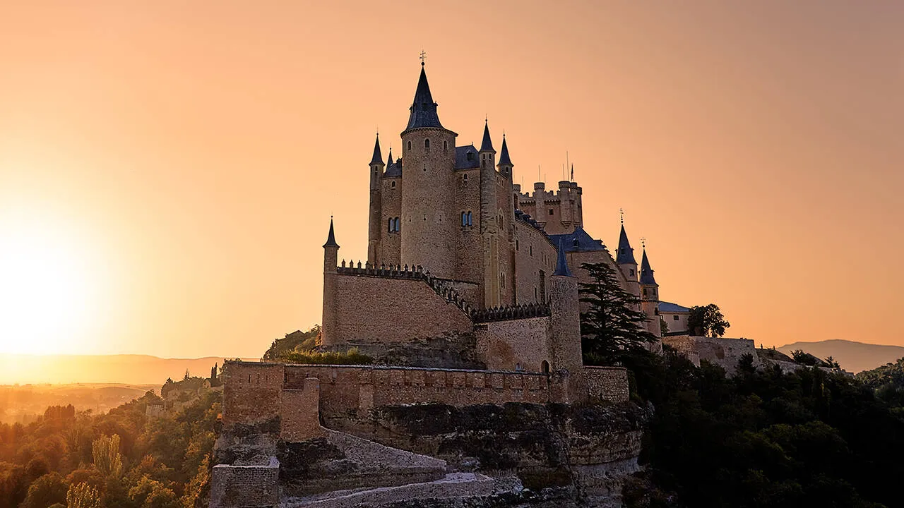 The Alcázar of Segovia on its rocky spur at sunset: slate-capped conical turrets and a tall square tower above crenellated walls, the sky glowing orange and the sun low on the left