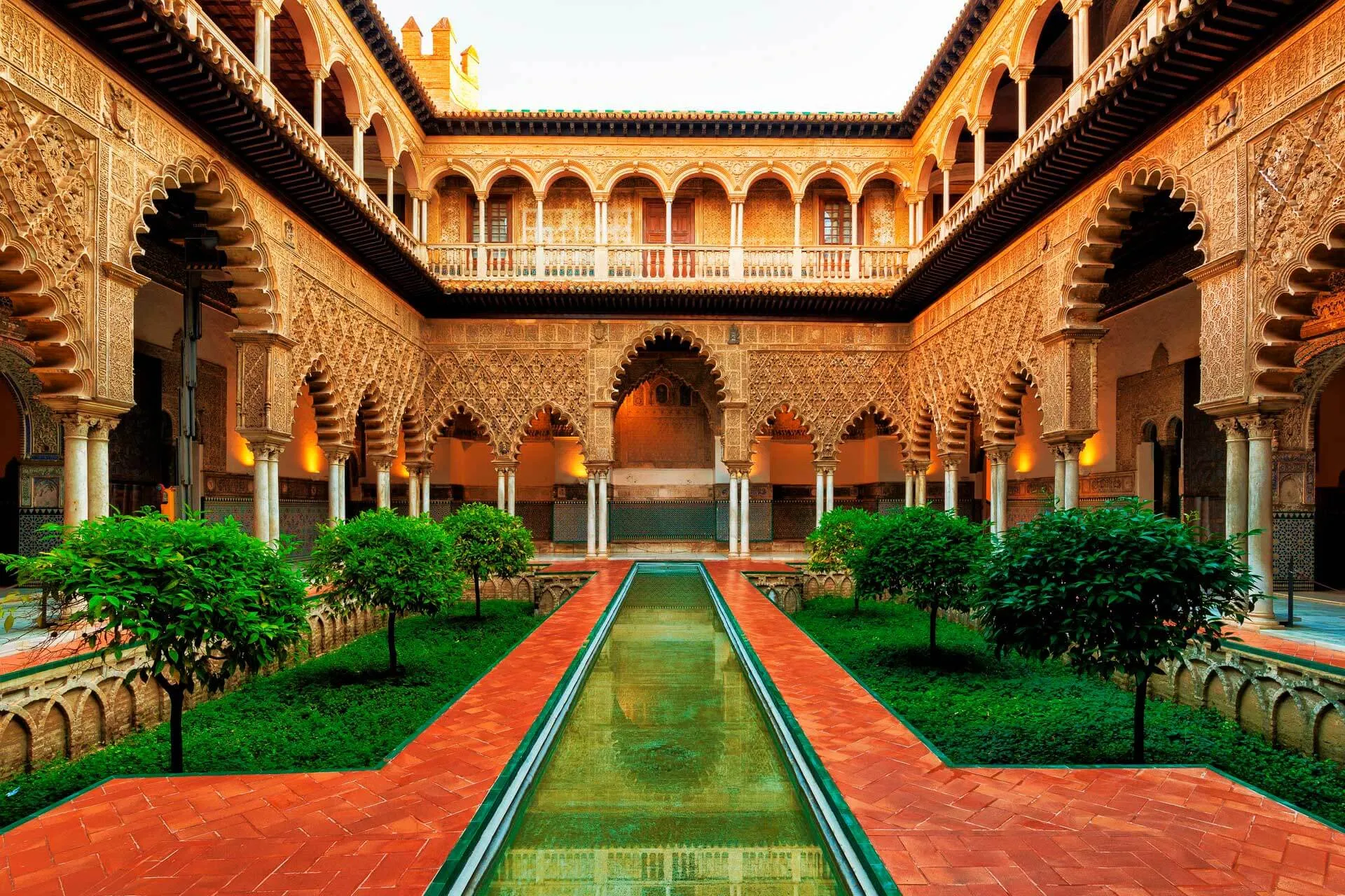 The Patio de las Doncellas in Seville's Real Alcázar: carved, many-lobed arches on marble columns around the courtyard, lamplight in the arcades and a gallery above, a long narrow pool running between sunken gardens of small trees and a brick floor