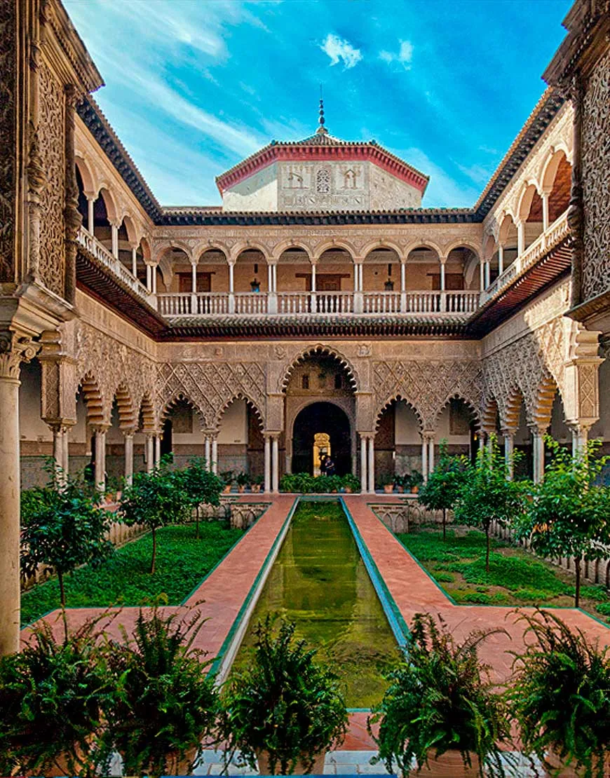 A courtyard of the Real Alcázar: a long, narrow pool between sunken gardens, framed by carved, lobed arches, with a balustraded gallery above and a tiled pavilion roof beyond under a blue sky