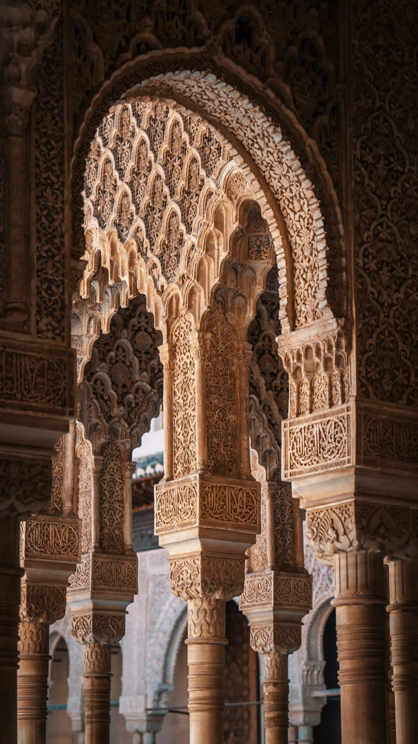 Carved stucco arches in the Alhambra: a pointed arch hung with honeycomb muqarnas above slender columns, further arcades receding behind it in warm light
