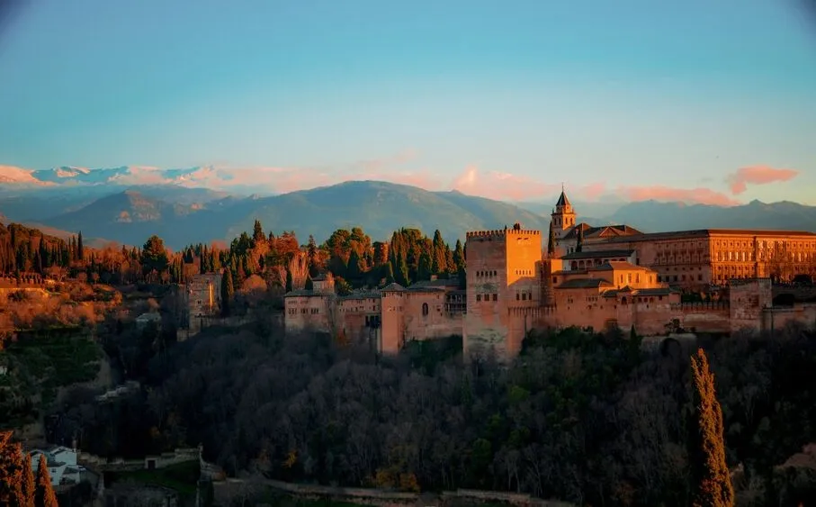 The Alhambra on its wooded hill in Granada, its towers and palace walls lit orange at sunset, with the snow-capped Sierra Nevada behind