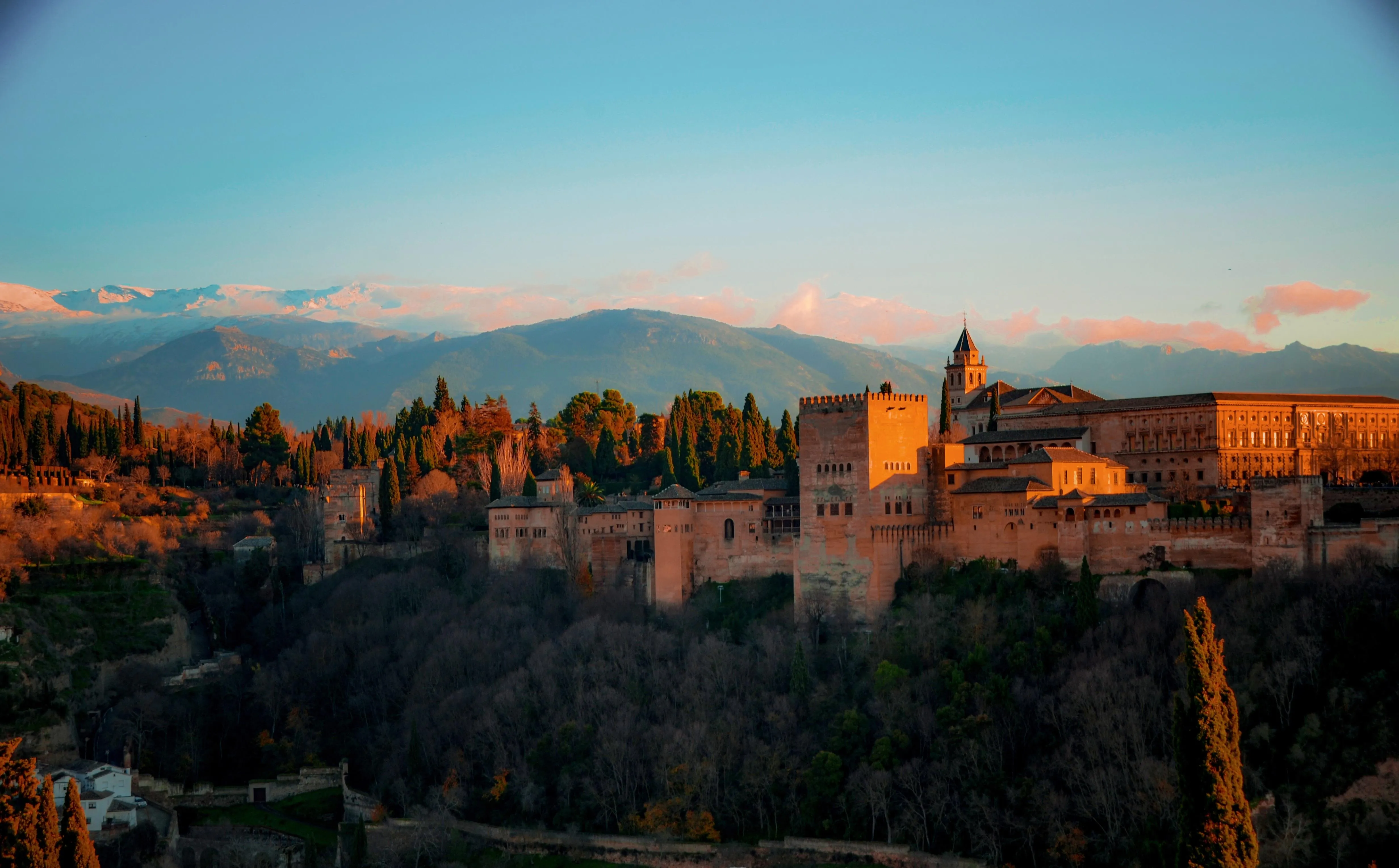 The Alhambra at sunset seen across the valley: its walls and square towers lit orange above dark woods, the snow-tipped ridge of the Sierra Nevada behind under a pink-streaked sky