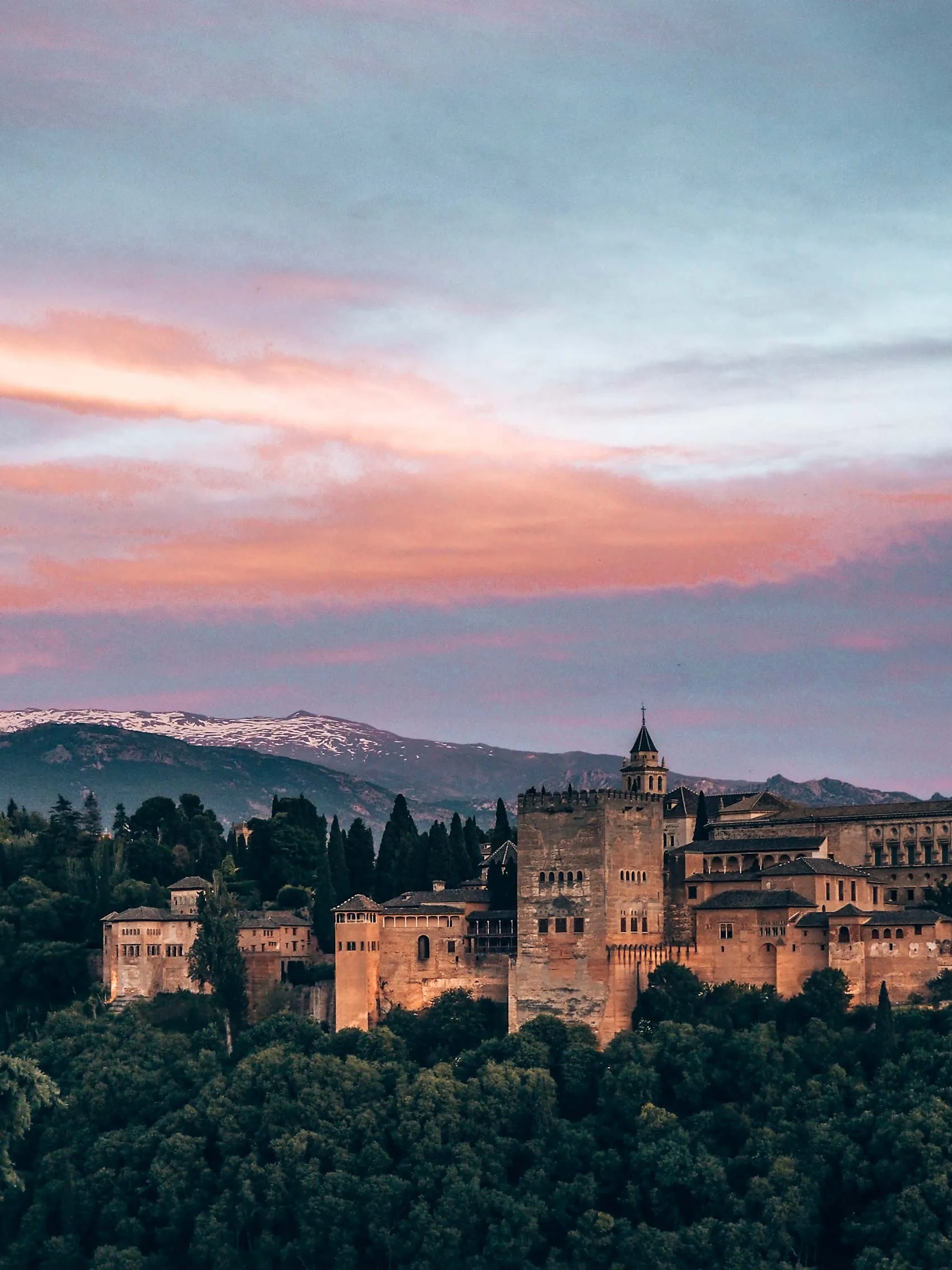The Alhambra along its wooded ridge at dusk, under a pink sky, with the snow on the Sierra Nevada behind it