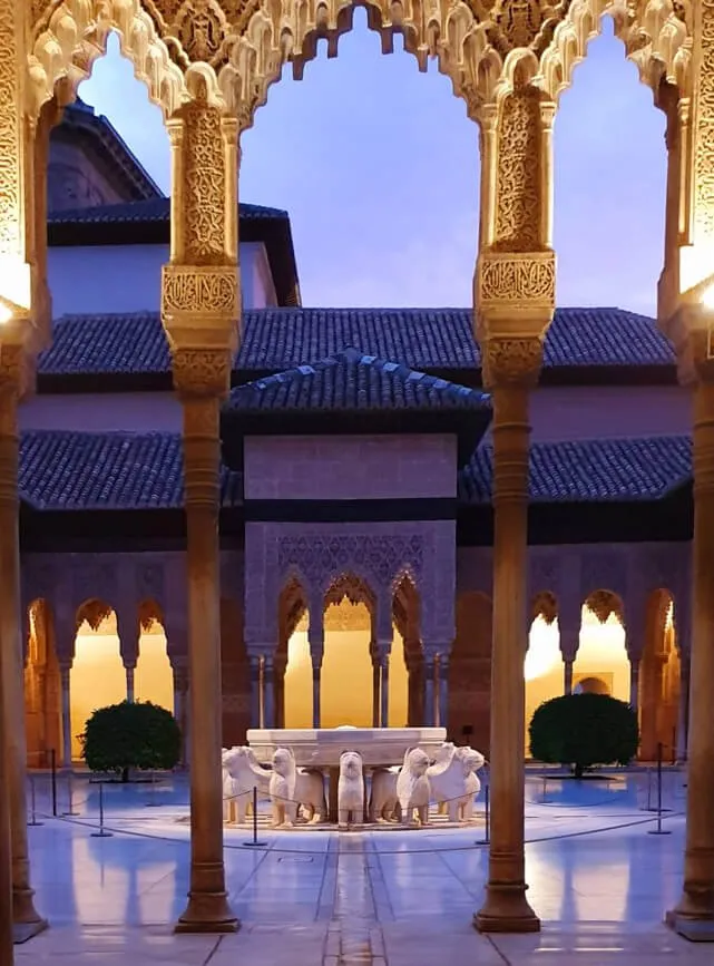 A courtyard seen through a carved stone arch at dusk: slender marble columns and lit arcades around a fountain ringed by stone lions, a tiled roof above