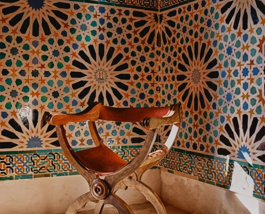 A wooden folding chair with a leather seat set in a corner of walls tiled in interlacing star patterns of blue, green, black and ochre