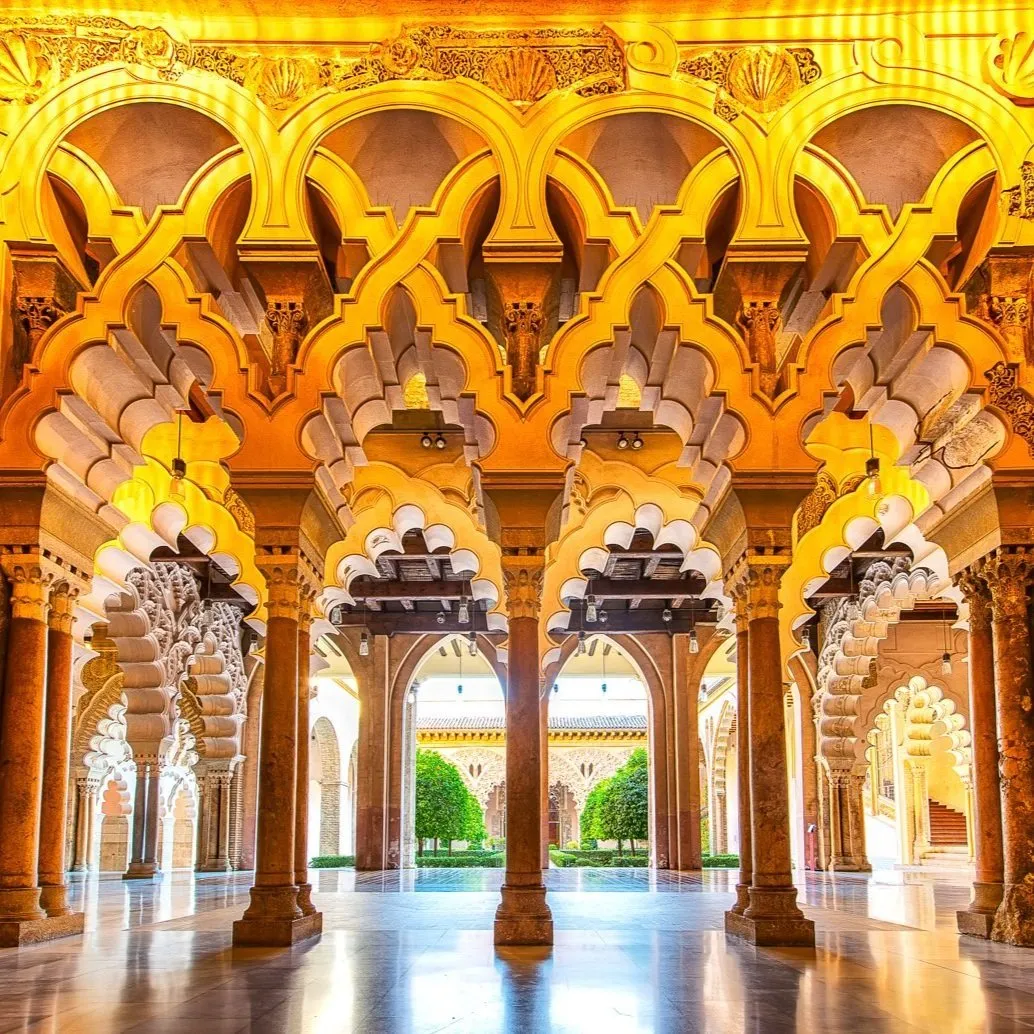 Interlaced, many-lobed arches lit gold in the Aljafería, carved plaster above slender columns, opening onto a courtyard garden of small trees beyond a polished floor