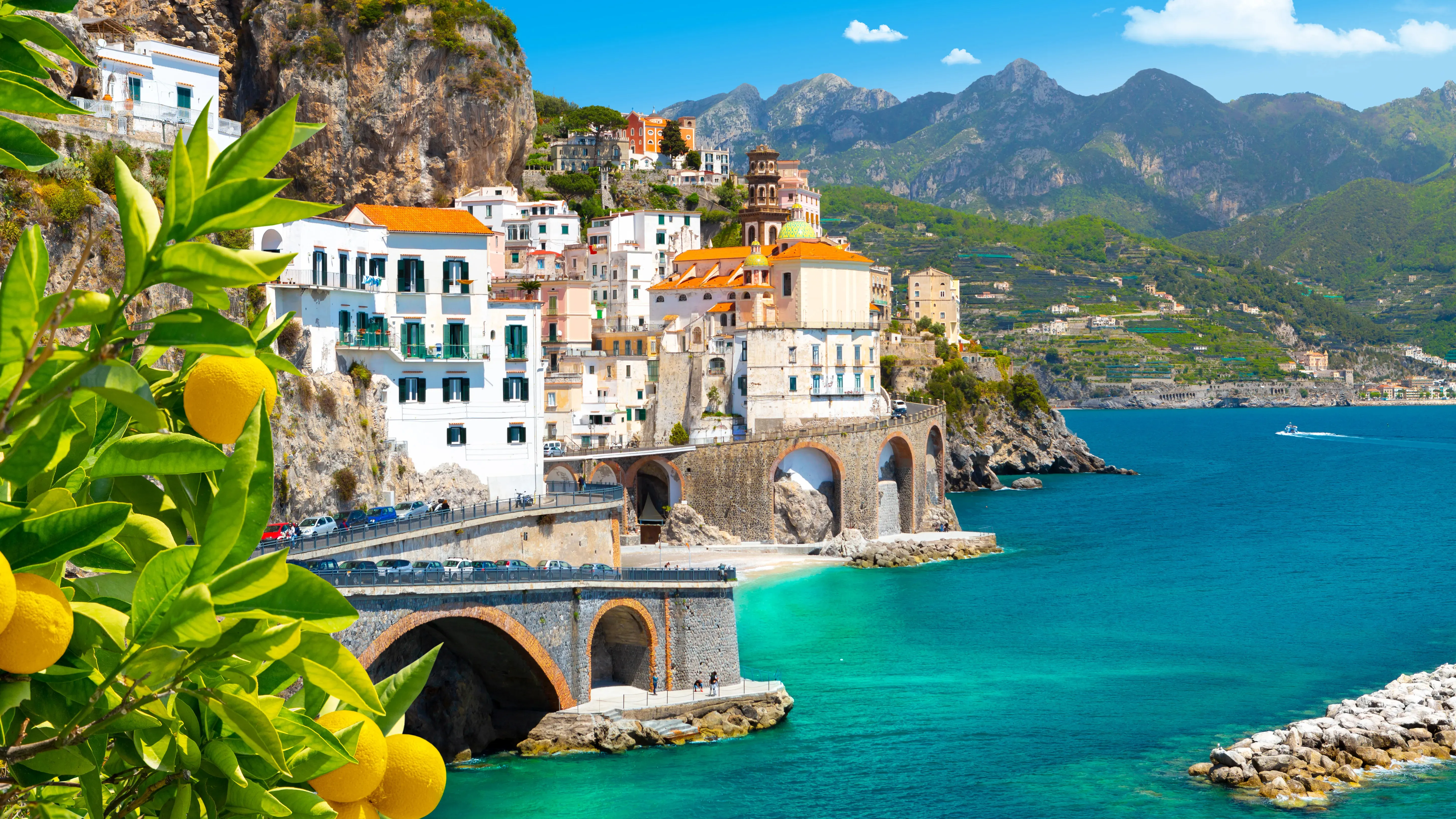 A small town of white and pastel houses on a cliff above a turquoise bay, a road crossing on stone arches below it, a bell tower with a tiled dome among the roofs and green mountains behind, with lemons on a branch in the foreground