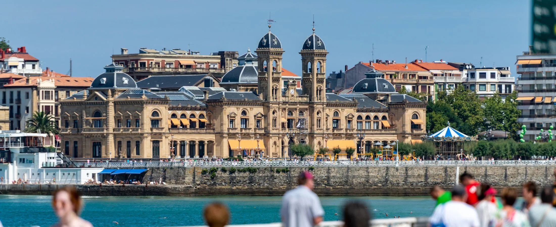 San Sebastián's City Hall across the water of La Concha: a long sandstone building with twin domed towers, slate roofs and orange awnings above the sea wall, a striped carousel among the trees at the right, and passers-by out of focus in the foreground