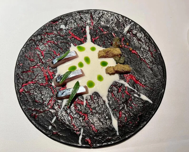A dish on a black plate veined in red like cooling lava: strips of silver-skinned fish with green leaves on a pool of white sauce dotted with green, and two crumbed pieces beside them