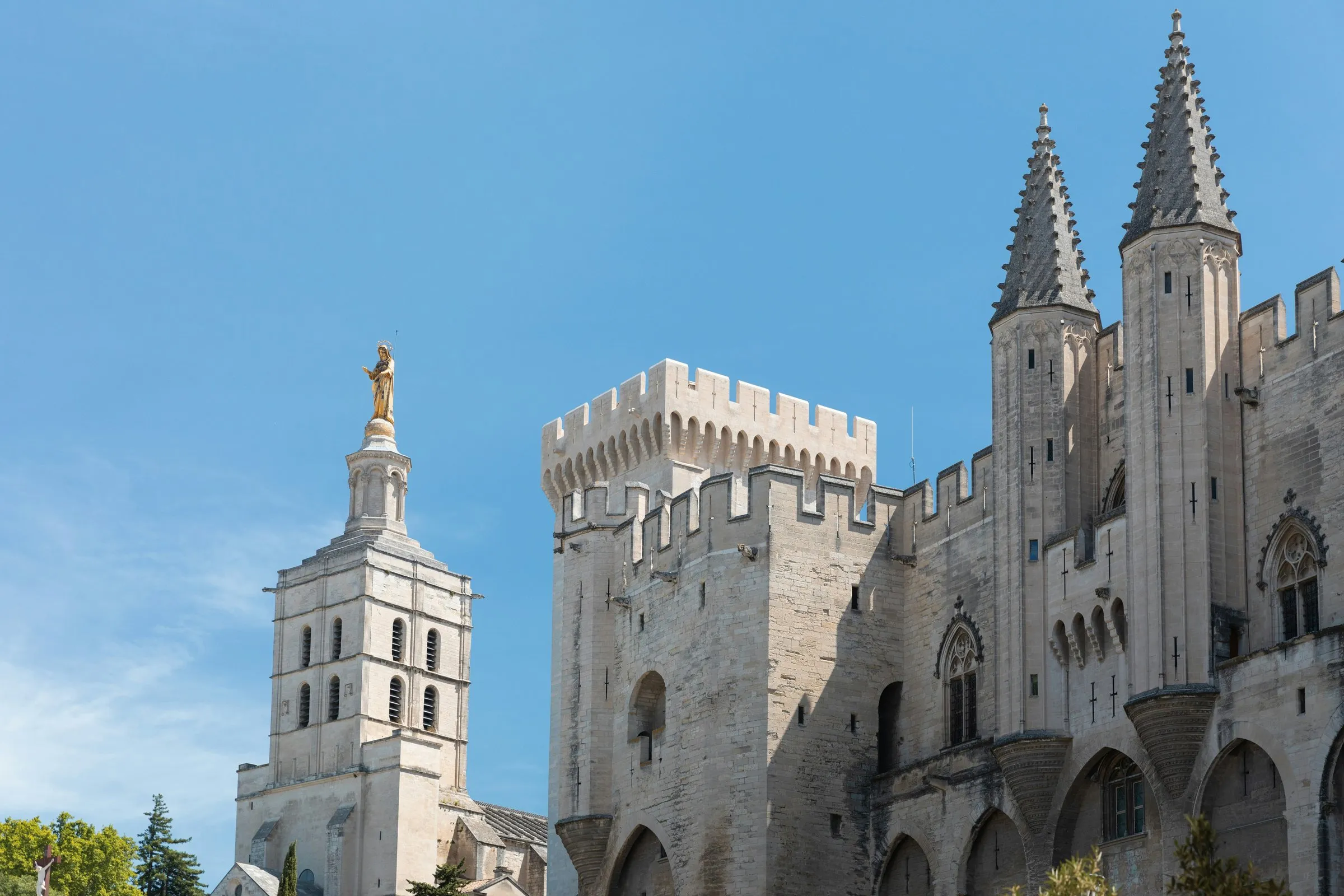 The Palace of the Popes in Avignon from the square before it: the gilded statue of the Virgin on the cathedral tower at the left, the crenellated Tour de la Campane, and the pale stone façade of the Palais Neuf with its two pointed turrets over the entrance gate, under a clear blue sky