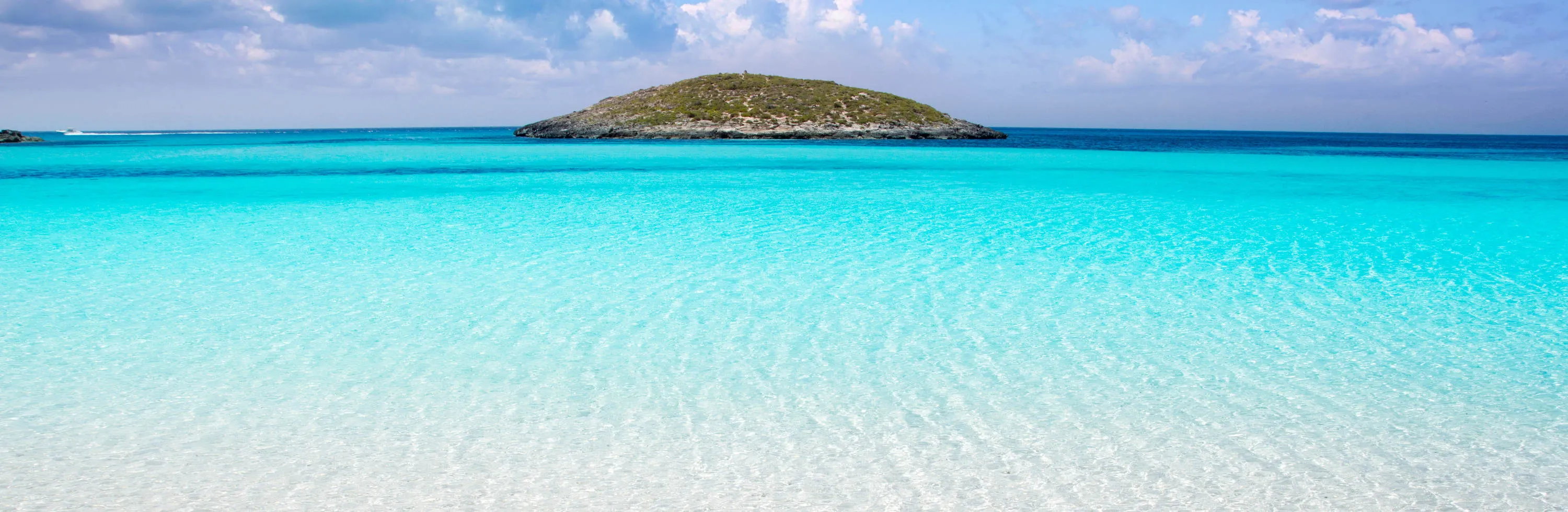 Clear turquoise shallows over pale sand, deepening to blue around a low, scrub-covered islet under a summer sky