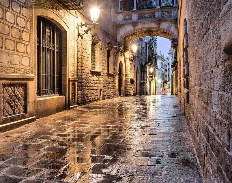 A narrow lane of wet flagstones between old stone buildings at dusk, wall lamps lit, under a carved stone bridge that joins the two sides of the street high above
