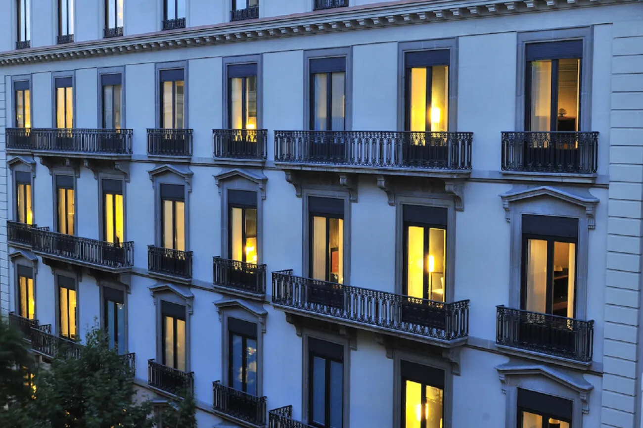 The hotel's pale stone façade at dusk: rows of tall windows lit gold behind wrought-iron balconies, with the top of a street tree in the lower corner