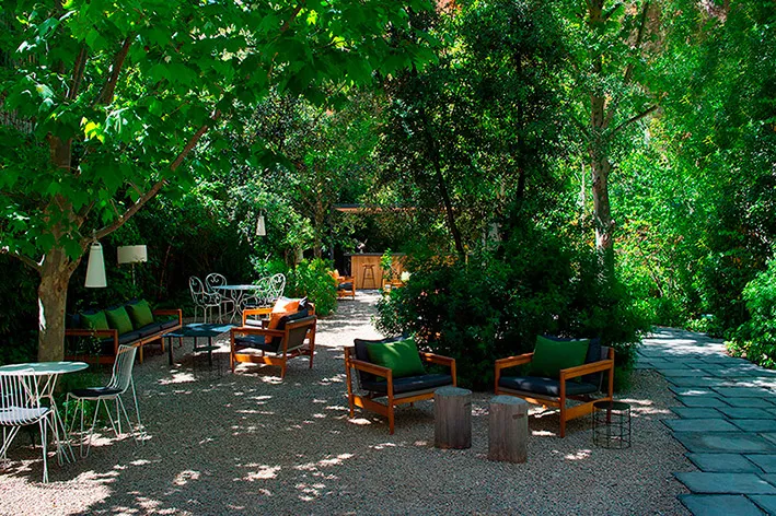 The hotel garden: armchairs and sofas with green cushions set out on gravel under tall trees, beside a slate path