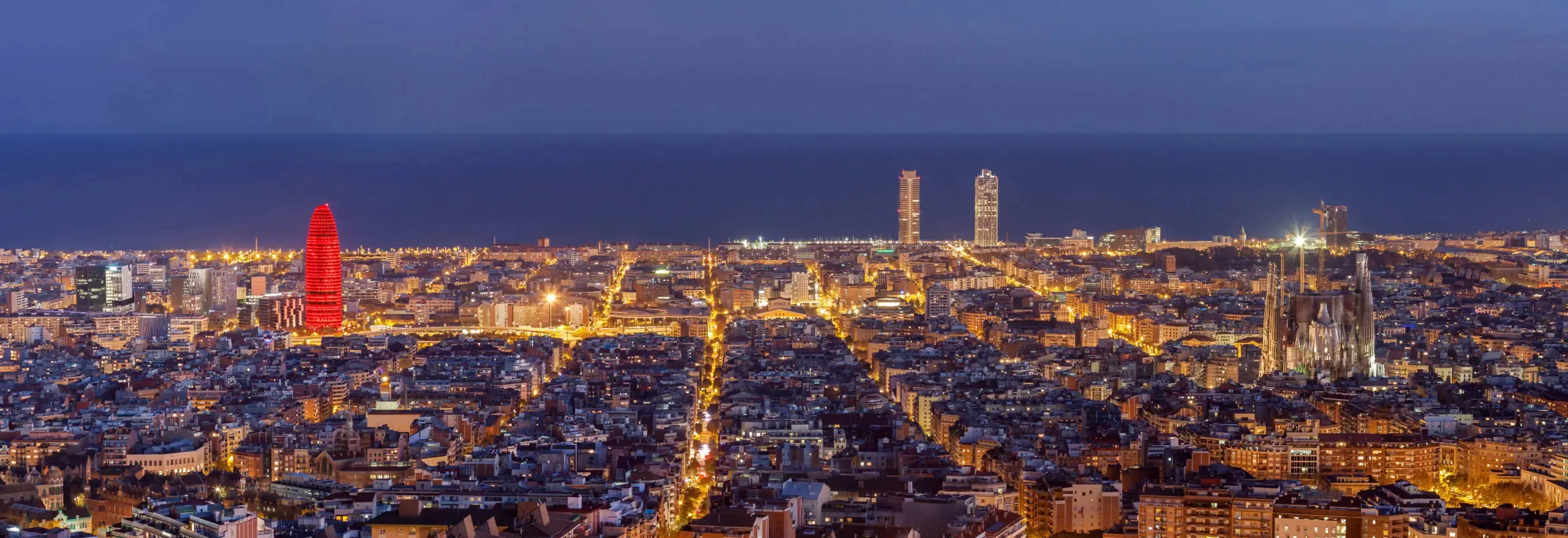 Barcelona at night from the hills: the lit grid of the Eixample running down to the sea, a bullet-shaped tower glowing red on the left, two tall towers on the waterfront ahead and the Sagrada Família on the right