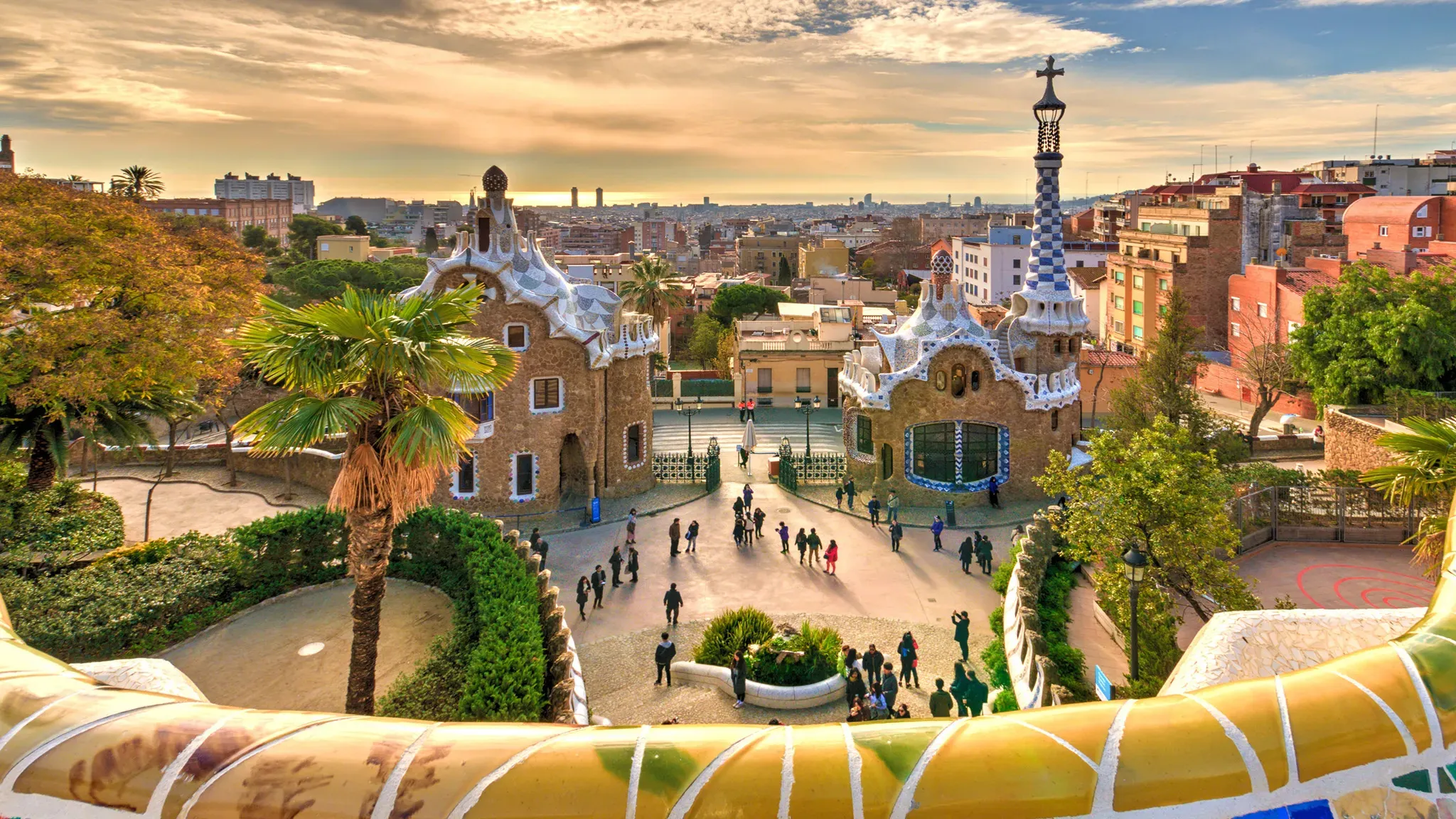 Park Güell from its mosaic bench: two gingerbread-like gatehouses with patterned roofs and a tall spire, a square of small figures below and the city running to the sea