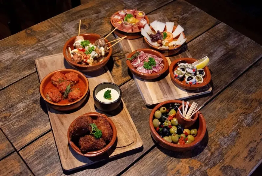 A tapas table from above: small terracotta dishes of croquetas, olives, cured meats, cheese and skewers on wooden boards, on a dark weathered table