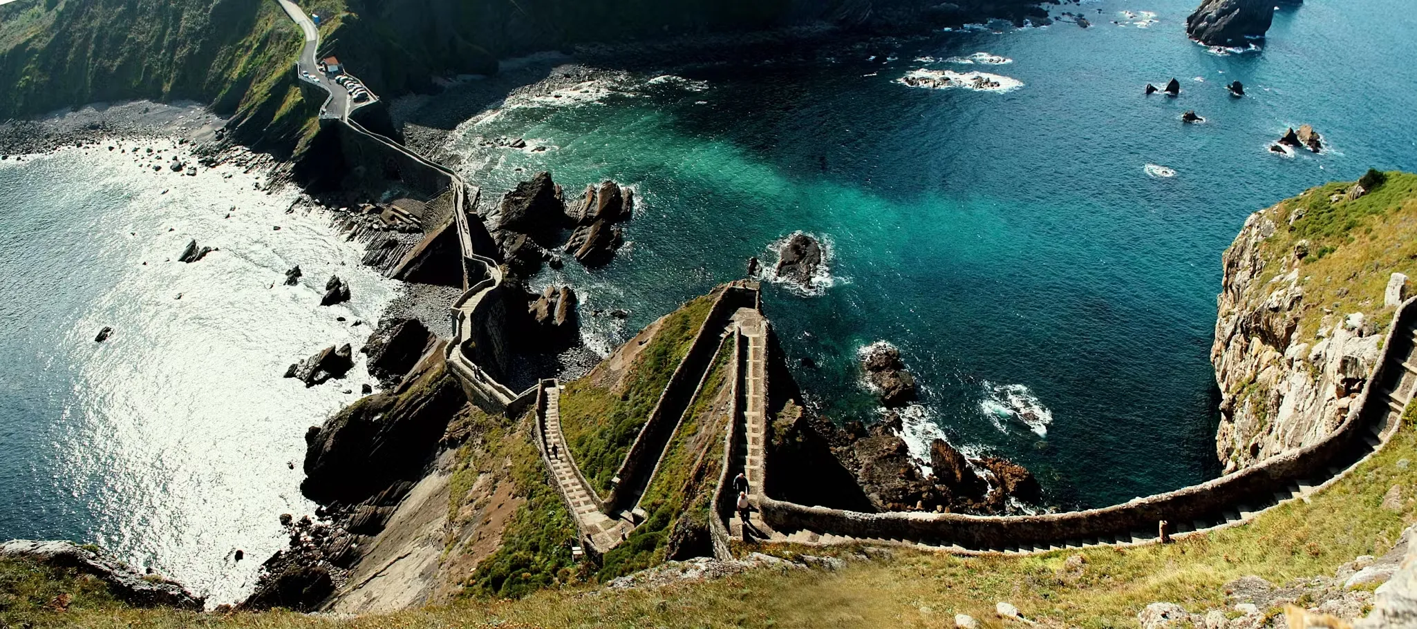 San Juan de Gaztelugatxe from high on the islet: stone steps zigzagging down to the bridge, the path running on along the cliffs to the mainland, and a green-blue sea breaking on the rocks