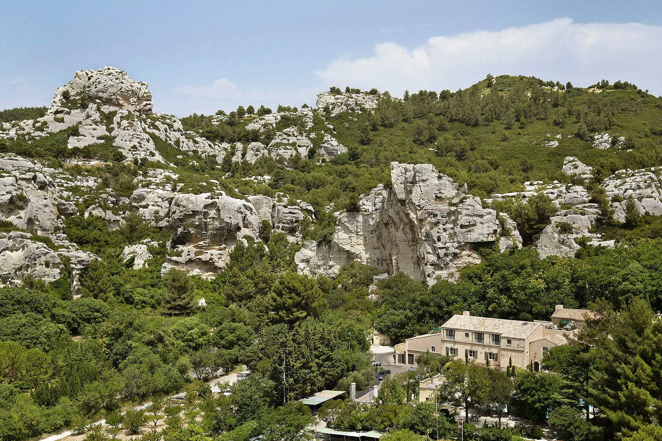 The hotel's main house, L'Oustau de Baumanière — an ochre two-storey mas with a tiled roof and pale shutters — seen from above on the green floor of the valley, pines and cypresses around it, a drive and a few parked cars at lower left, the white limestone cra