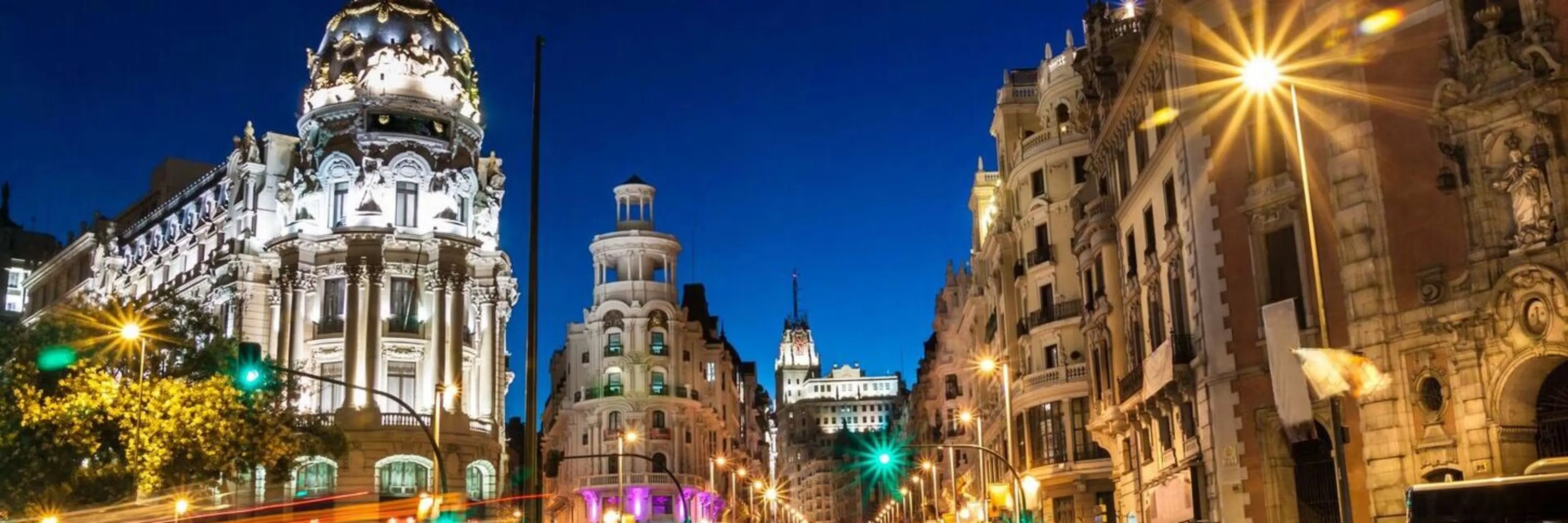 The Gran Vía at dusk under a deep blue sky: the floodlit dome of the Metropolis building on the left, the turreted Edificio Grassy ahead, street lamps flaring and traffic lights glowing green down the street