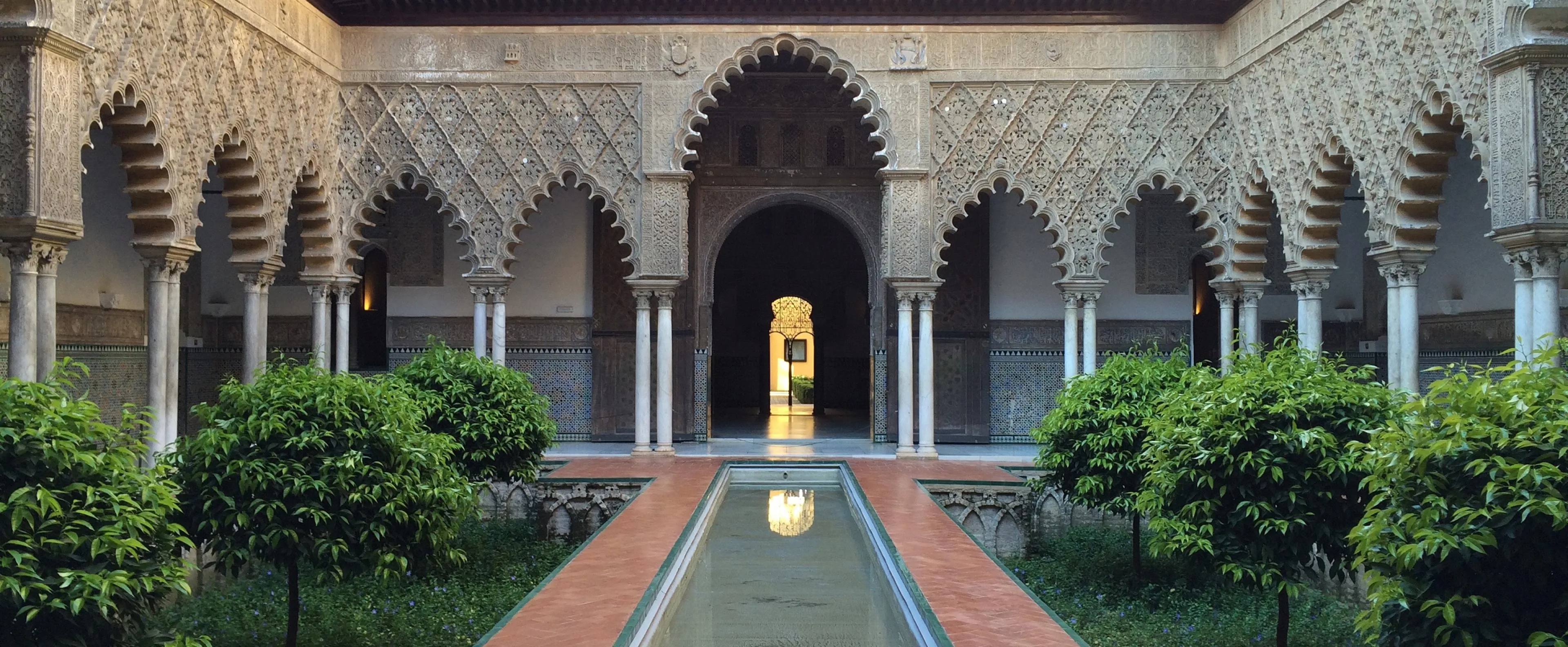The Patio de las Doncellas in the Real Alcázar: a long, narrow pool between sunken gardens of small trees, under scalloped arches of carved stucco on paired marble columns