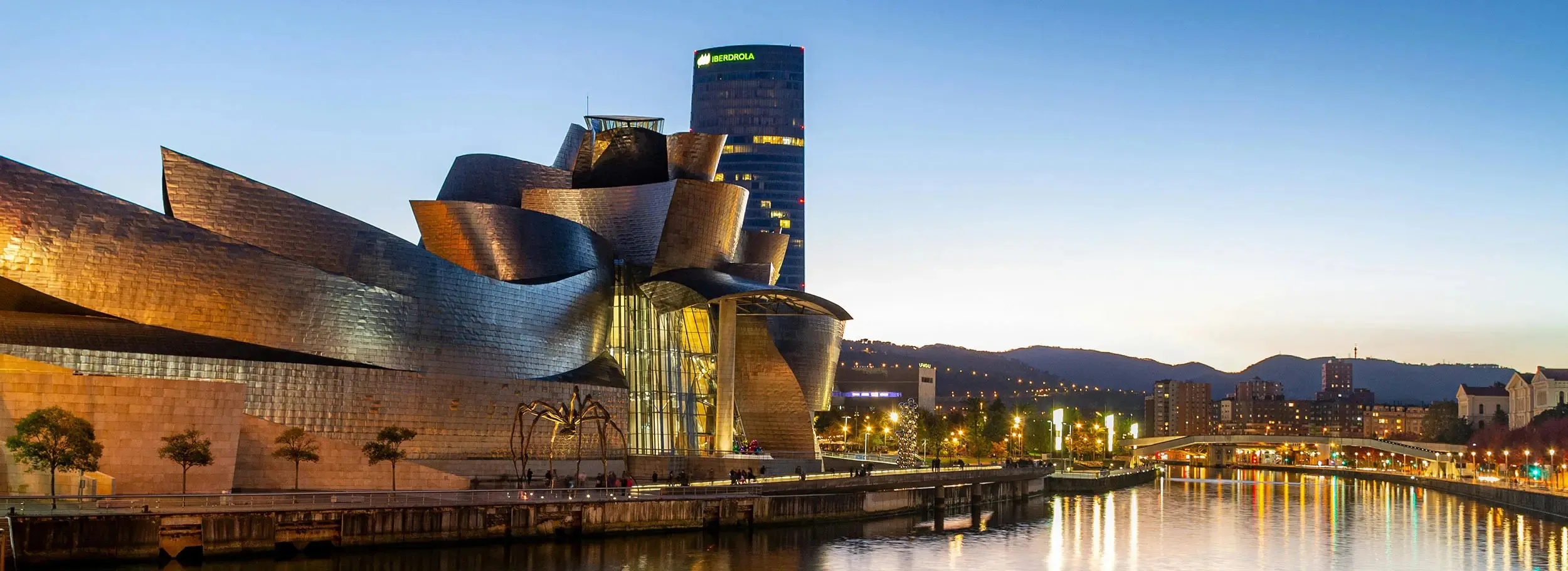 The Guggenheim Museum Bilbao at dusk from the riverside, its curved metal forms lit gold, a giant bronze spider on the walkway beside it, the Iberdrola Tower rising behind with its lit sign, and the lights of the far bank reflected in the river