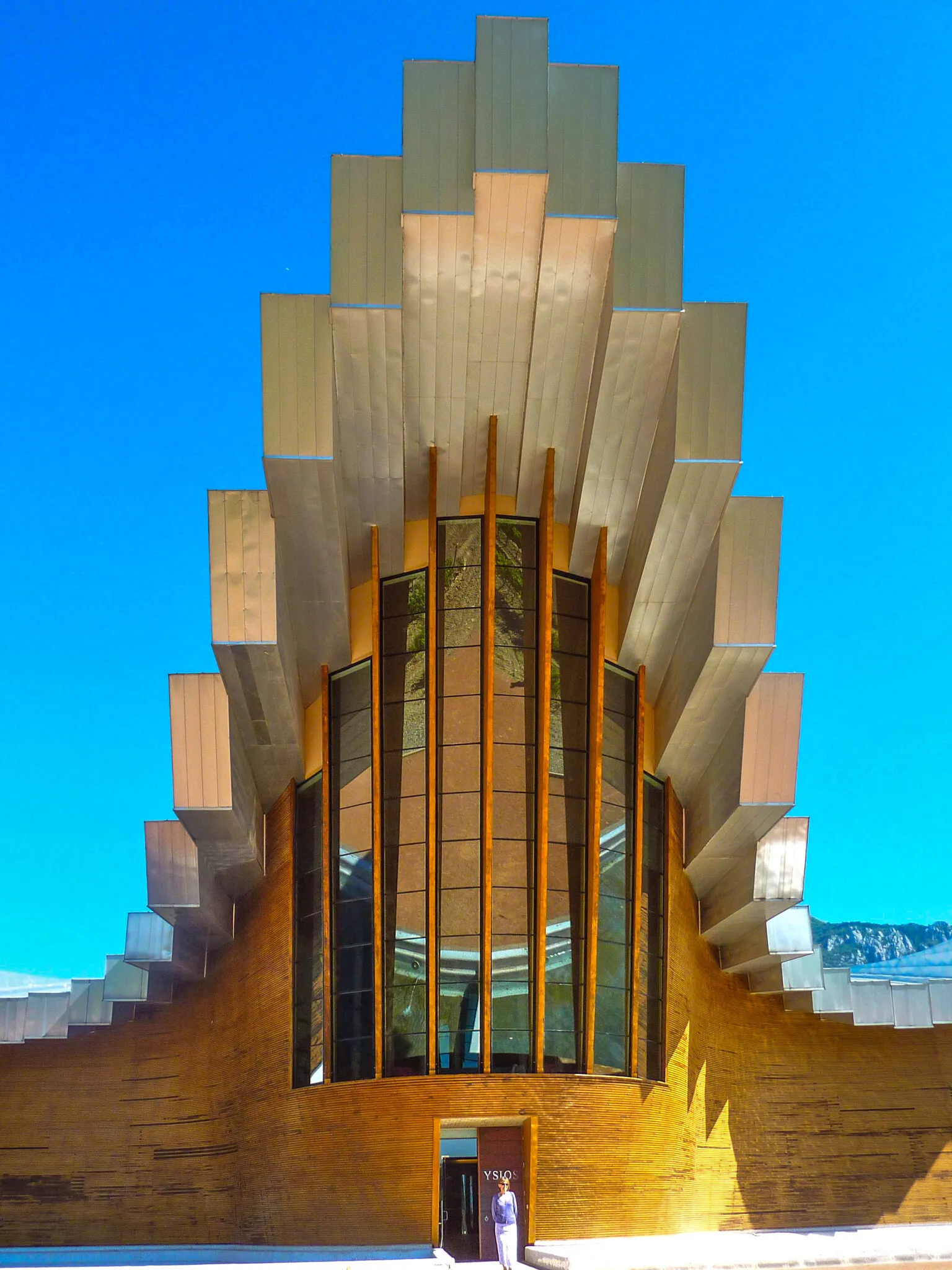The entrance front of Bodegas Ysios: stepped metal-clad roof panels rising in a crest above a tall window ribbed with timber, a curved wood-clad wall below with the name YSIOS on the door, under a deep blue sky