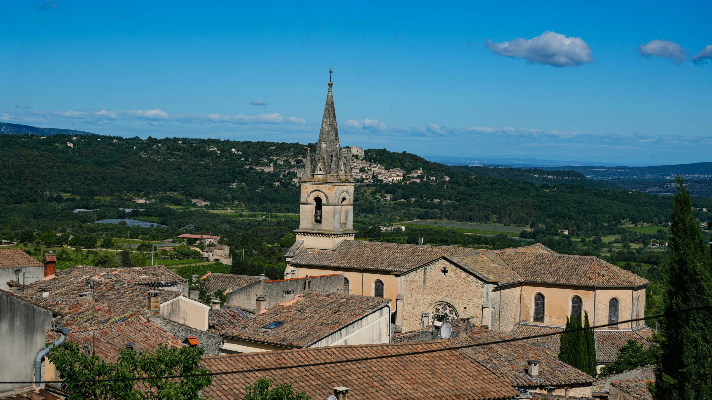 Bonnieux from the upper village: tiled roofs stepping down to the Église Neuve with its slender spire, the plain of the Calavon beyond, and the hill village of Lacoste with its château ruin on the facing slope under a blue sky