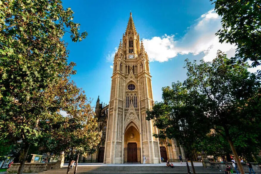 A tall neo-Gothic cathedral in pale stone, its single spired tower with a clock rising above the main door, seen from the steps of its square between trees under a blue sky