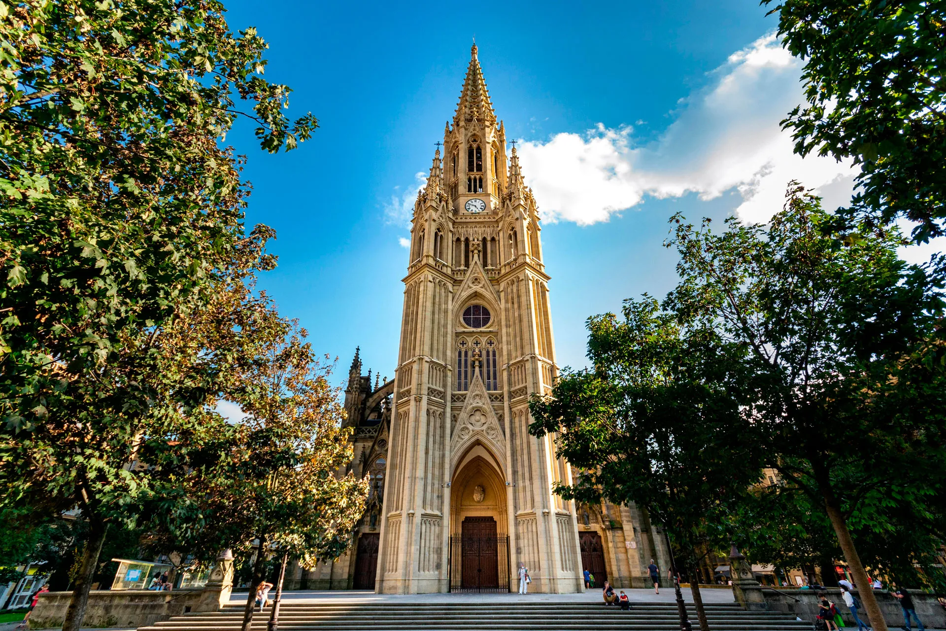 The neo-Gothic Buen Pastor Cathedral in San Sebastián, its tall clock tower and spire rising between trees against a blue sky, a wide flight of steps before its doors