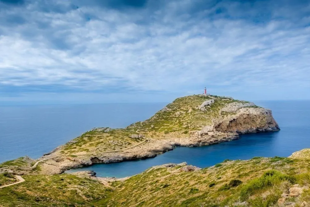 A rocky headland of scrub and pale limestone topped by a red-and-white lighthouse, above a sheltered deep-blue inlet, under a sky of broken cloud