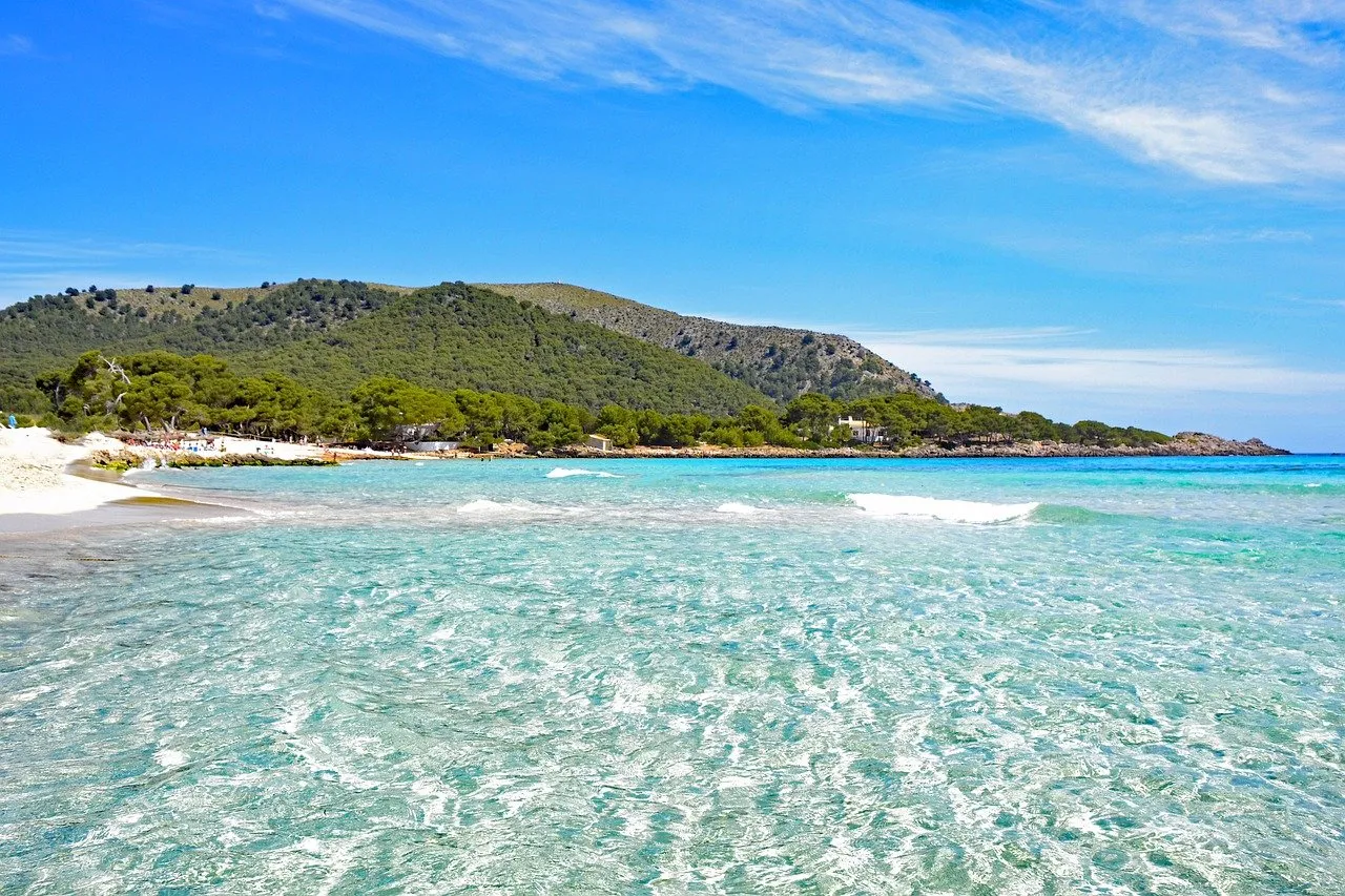 Clear turquoise water running up to a beach of white sand, low waves breaking, and hills of pine along the shore under a blue sky with thin cloud