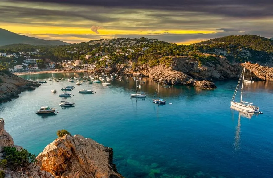 A sheltered bay of clear blue-green water with yachts at anchor, pine-covered slopes and white houses around it, under a golden evening sky