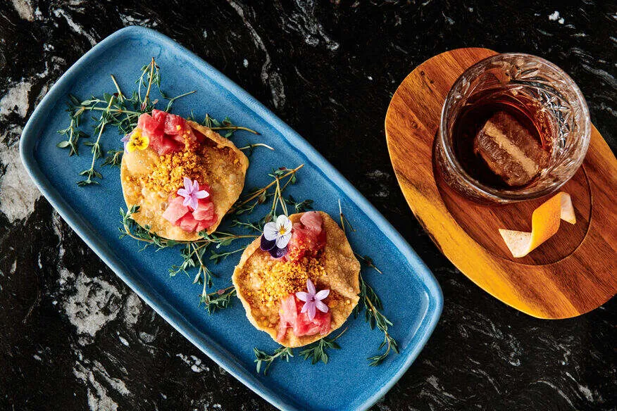 Two small tapas on a long blue plate, topped with pink slices, crumbs and tiny edible flowers, beside a tumbler of amber drink on a round wooden board, on dark marble