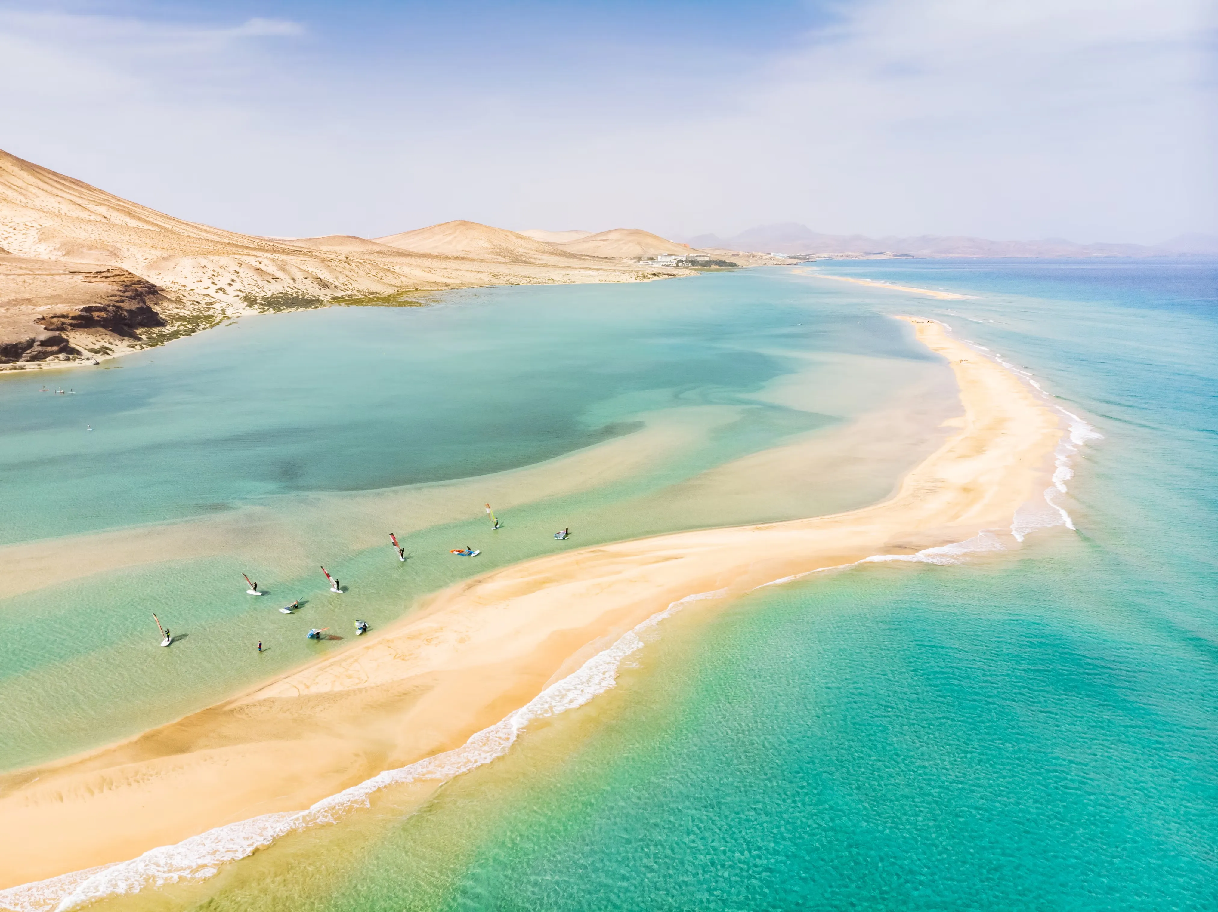 A curving sandbar at Playa de Sotavento de Jandía seen from above, with windsurfers in the shallow turquoise lagoon behind it