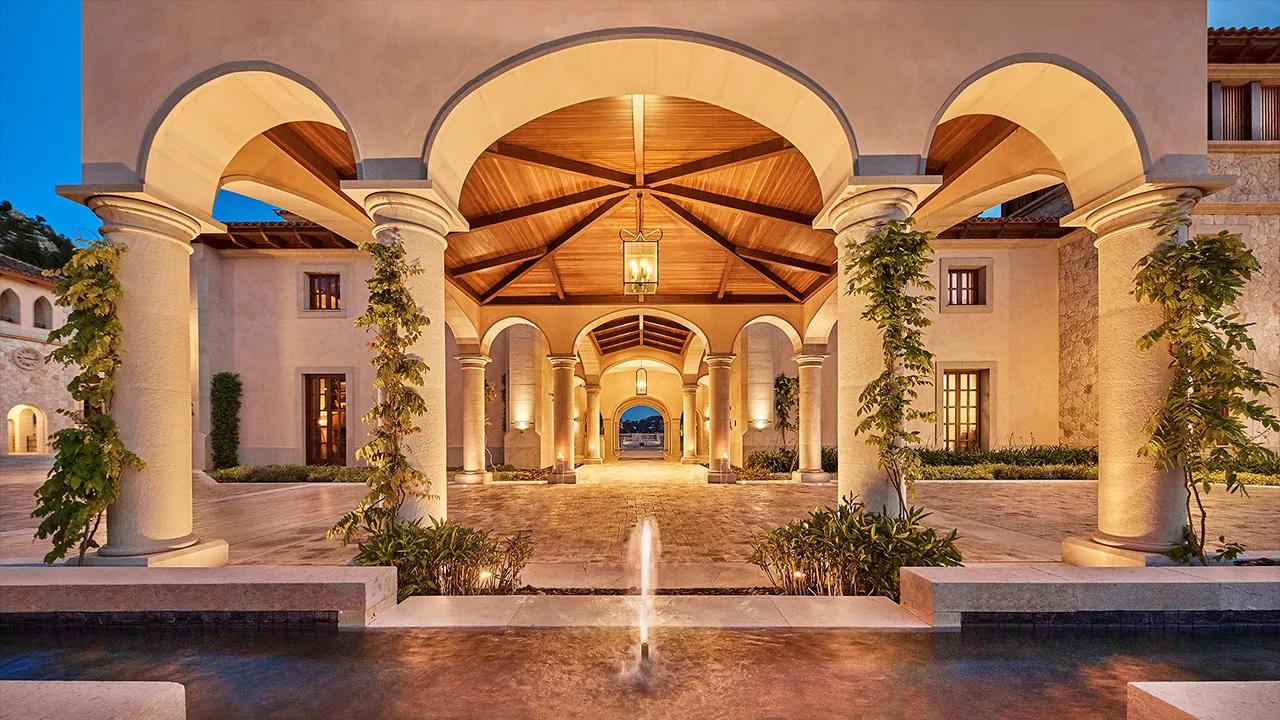 Arched entrance of Cap Vermell Grand Hotel at dusk, with a lantern-lit timber vault, climbing plants on the columns and a fountain in the reflecting pool