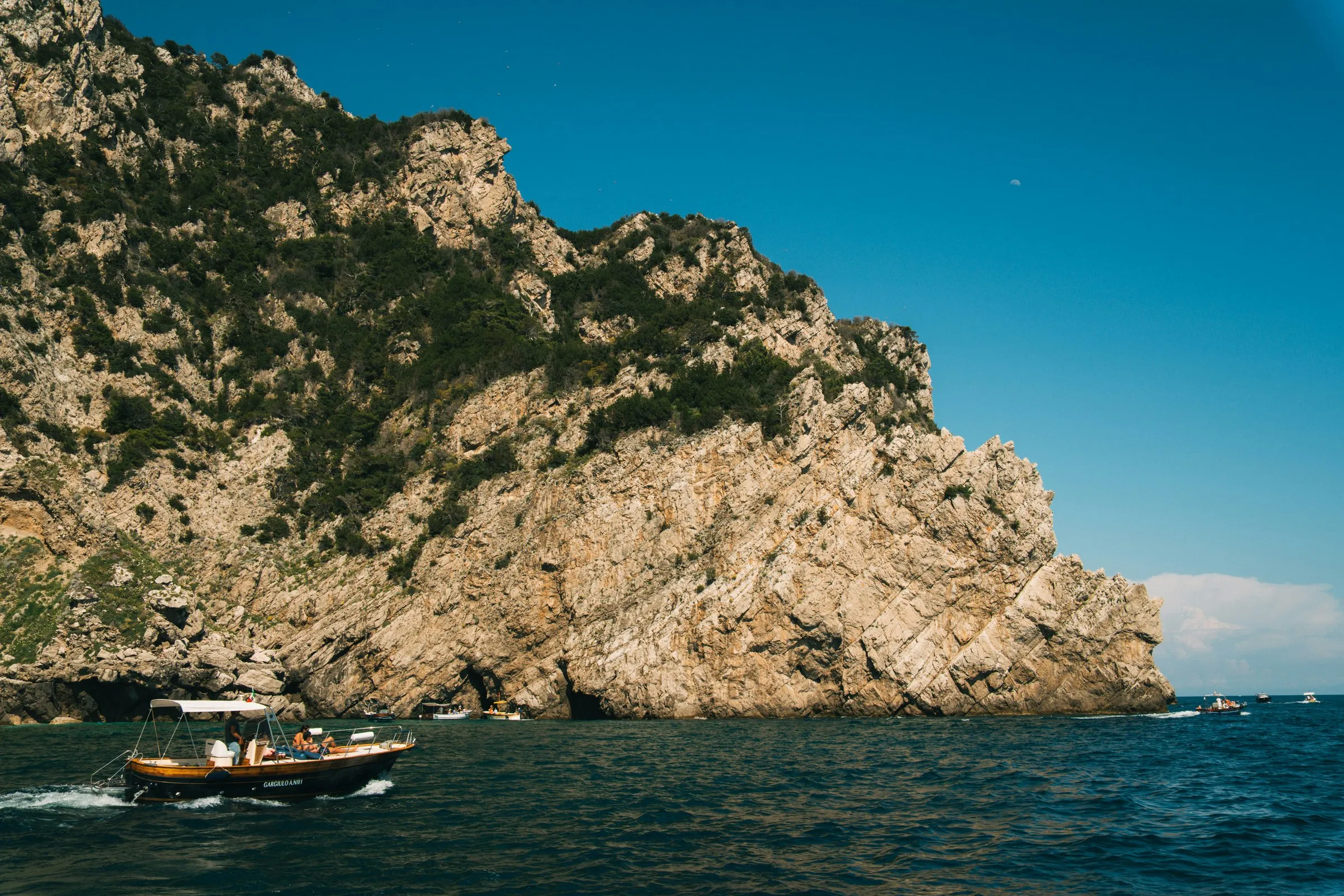 A wooden open boat with a white canopy under way along a limestone headland off Capri, sea caves at the waterline, a few small boats beyond and a deep blue sky