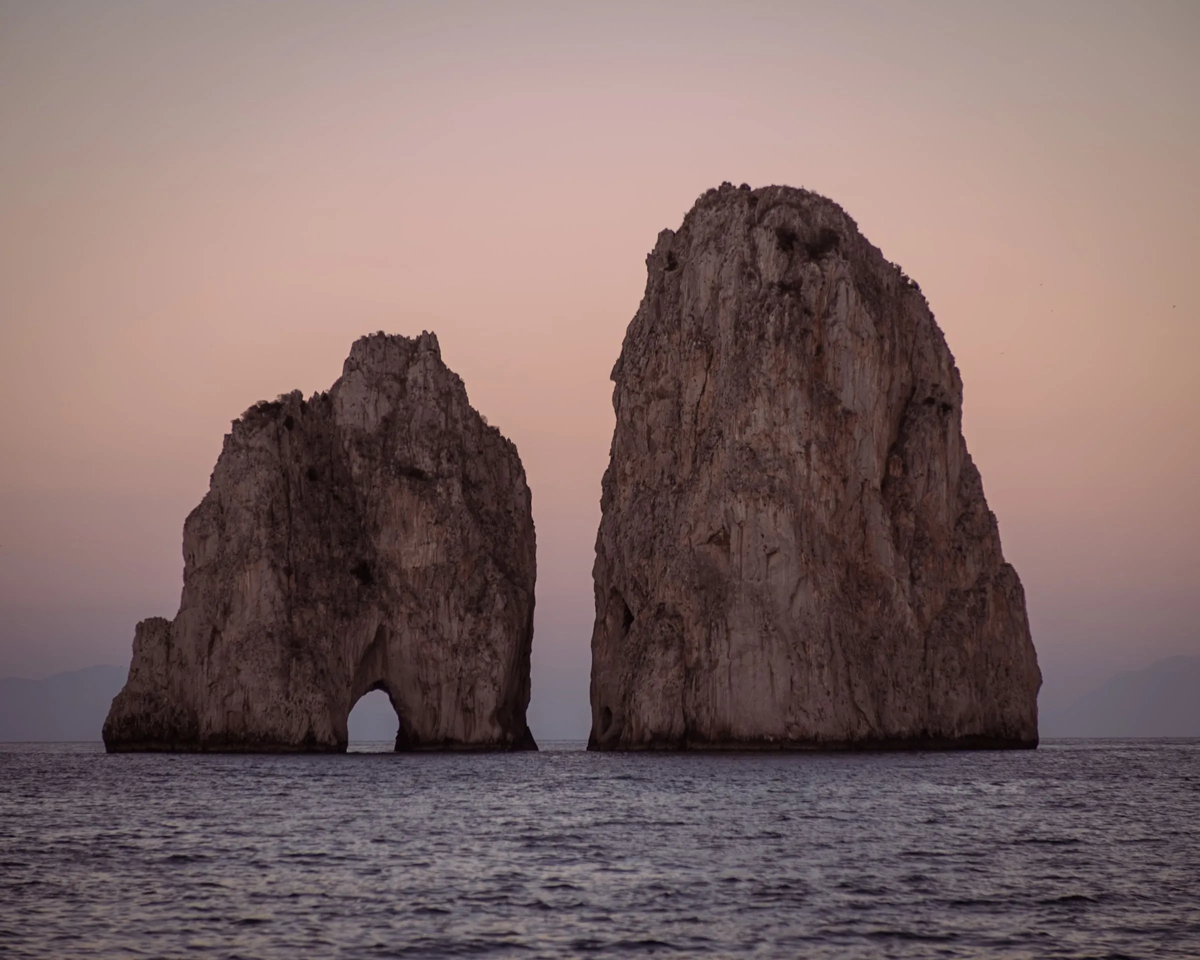 The Faraglioni off Capri at dusk: two great limestone stacks rising from a calm sea, the nearer one pierced by its arch, against a sky turned pink