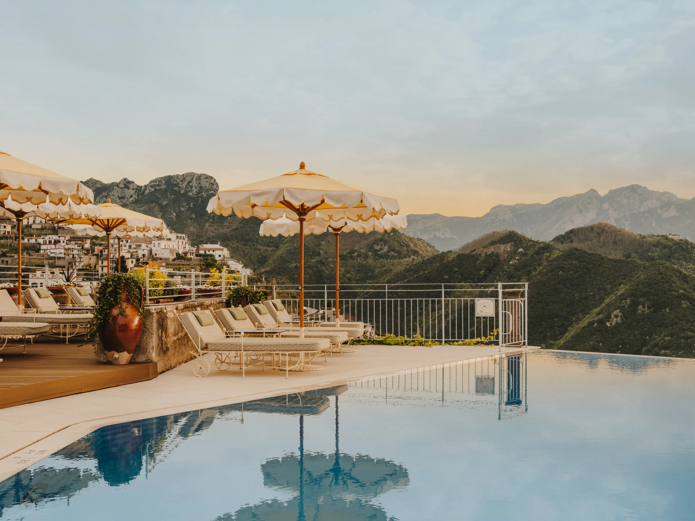 The infinity pool in soft golden light: fringed cream parasols on wooden poles over white-cushioned loungers on a stone-and-timber deck, a terracotta urn, a white iron railing along the far edge, the still water reflecting the parasols; the houses of Ravello o