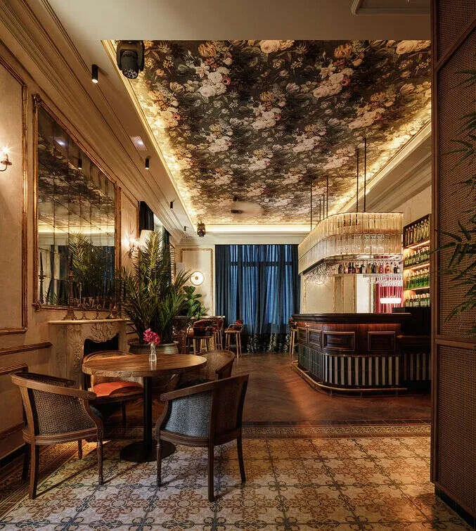 A bar and dining room under a ceiling papered with large flowers, with cane chairs at a small table on patterned floor tiles