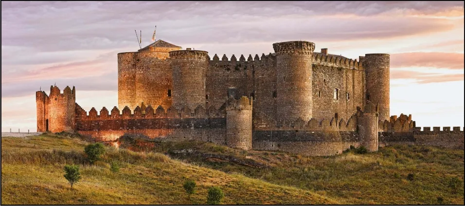 Belmonte Castle on a grassy hill in evening light: round towers and crenellated walls around a tall keep, an outer battlemented wall in front, under a pink and grey sky