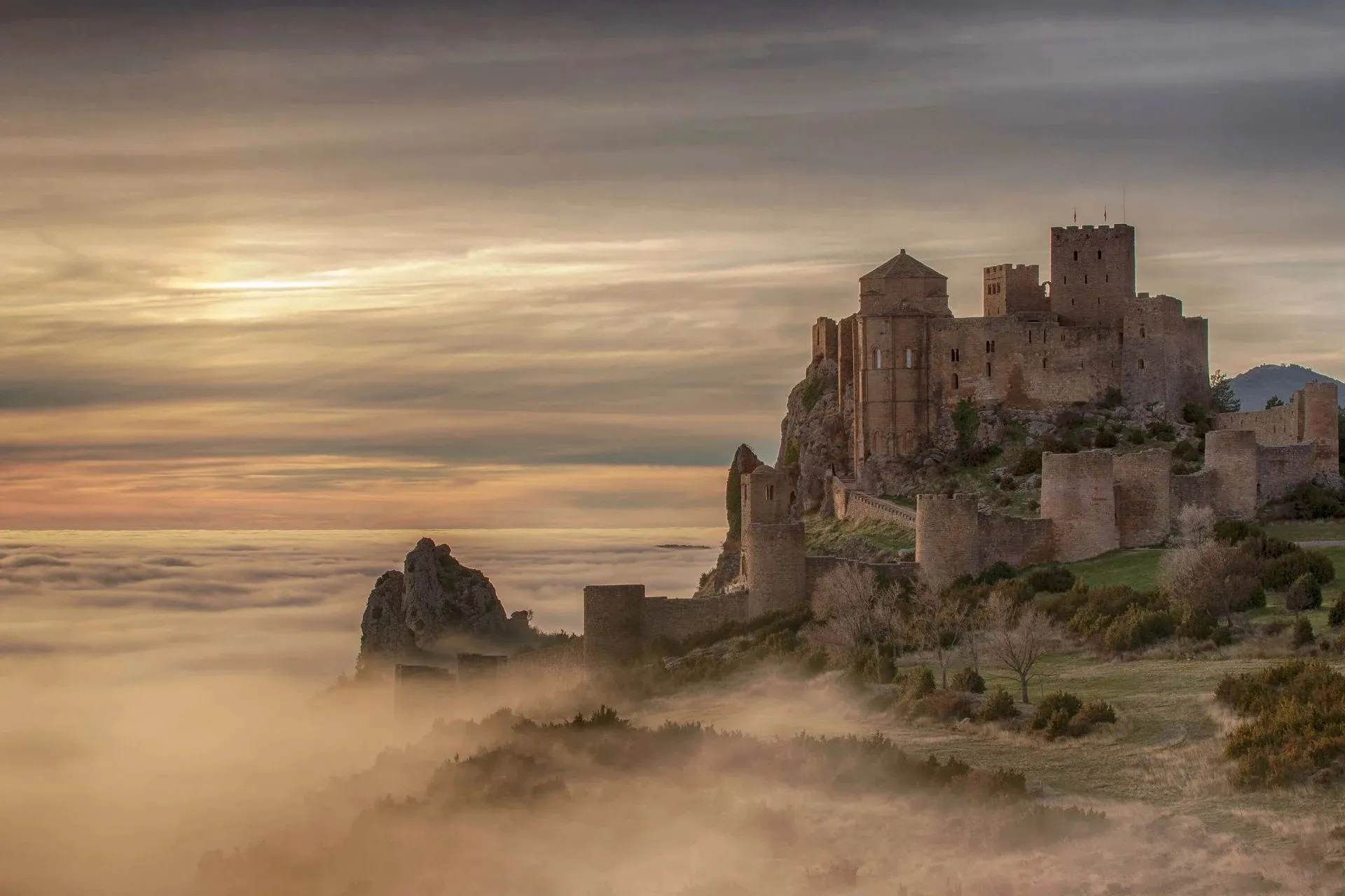 Loarre Castle on its rocky ridge in soft golden light, square towers and long walls rising above a sea of fog that fills the valley below