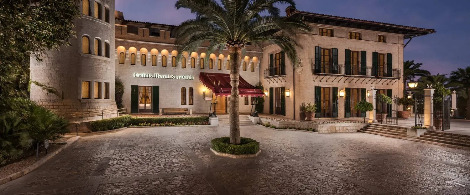 Entrance forecourt of Castillo Hotel Son Vida at dusk: a round stone tower, lit arched windows, a red awning over the door and a palm tree in the cobbled courtyard