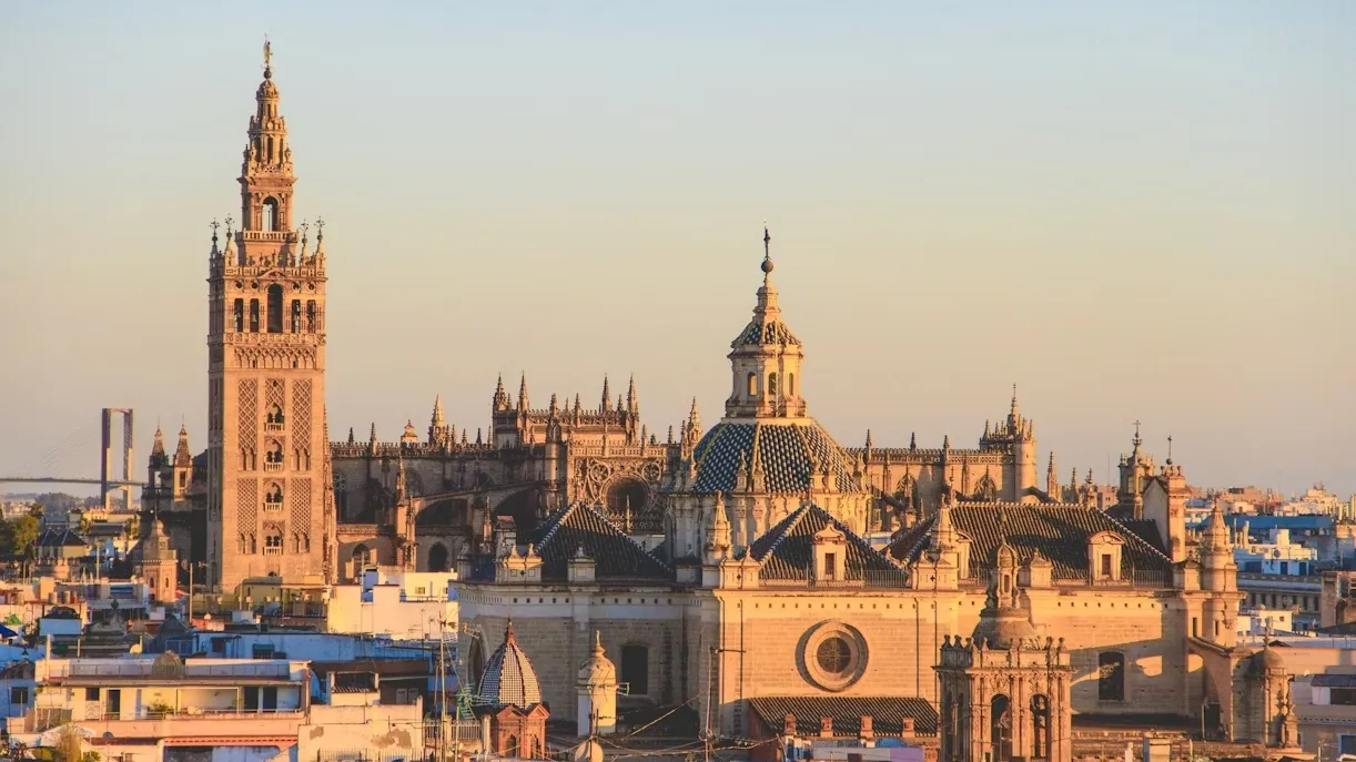 Seville Cathedral and the Giralda in warm evening light above the city's rooftops: the tall, ornate bell tower on the left, the Gothic pinnacles and a tiled dome beside it, a pale sky behind