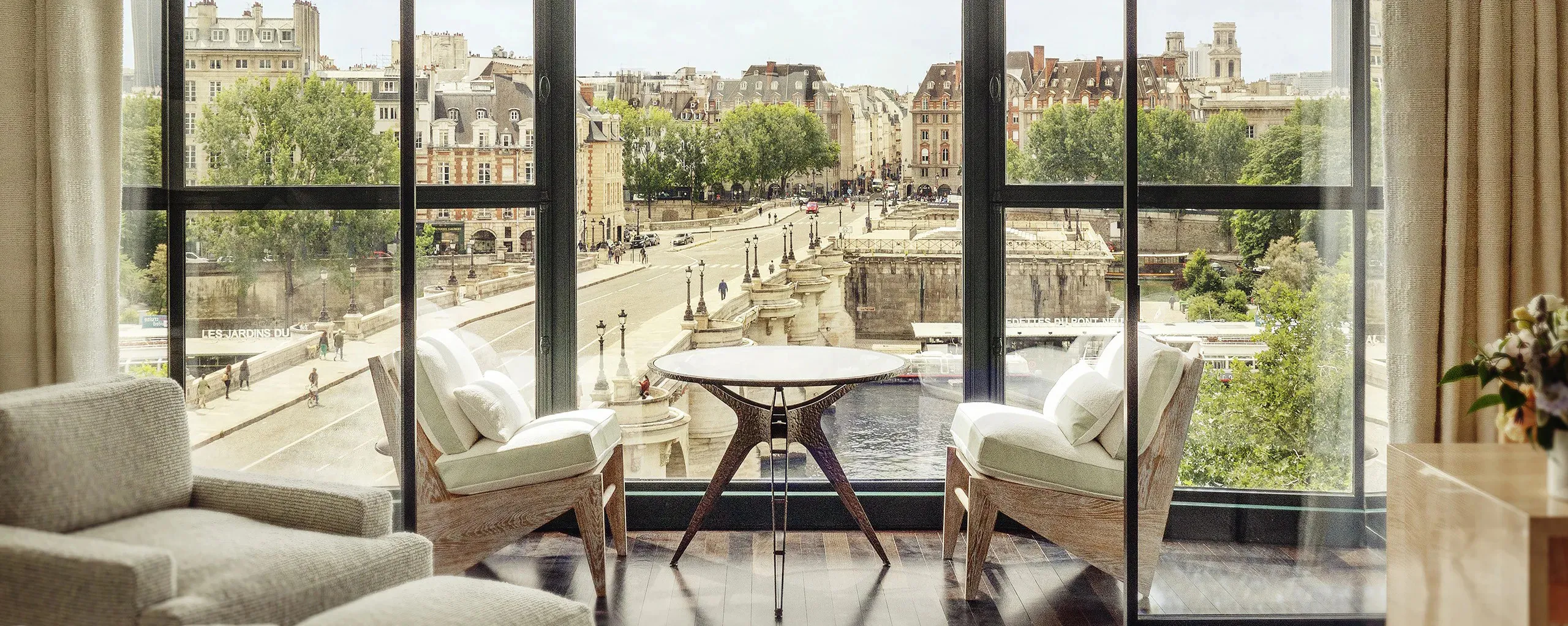 Two armchairs and a round table at a hotel room's tall windows over the Seine, and beyond the glass the Pont Neuf crossing to the tip of the Île de la Cité, its stone houses, trees and moored boats