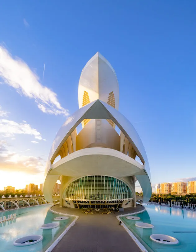 The Palau de les Arts seen head-on in late sun: a tall white shell curving to a point over a glass front, turquoise pools with round planters either side of a paved approach, and apartment blocks lit gold beyond