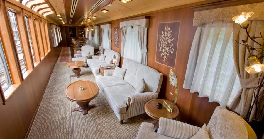 A railway saloon car panelled in light wood, with cream sofas and armchairs, round wooden side tables, net curtains at the windows and inlaid flower panels on the walls